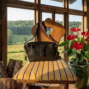 May include: A black leather crossbody bag with a buckle closure sits on a vintage chair with a striped cushion. The chair has a heart-shaped cutout in the back. A potted plant with red flowers is to the right, and a basket of logs is to the left.