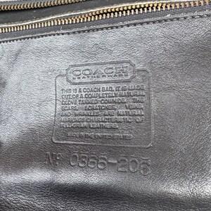 May include: Close-up of a black leather Coach bag. The bag has a textured surface with a gold zipper and a rectangular leather tag with text. The tag reads "Coach Leatherware" and includes text about the bag's materials and origin.