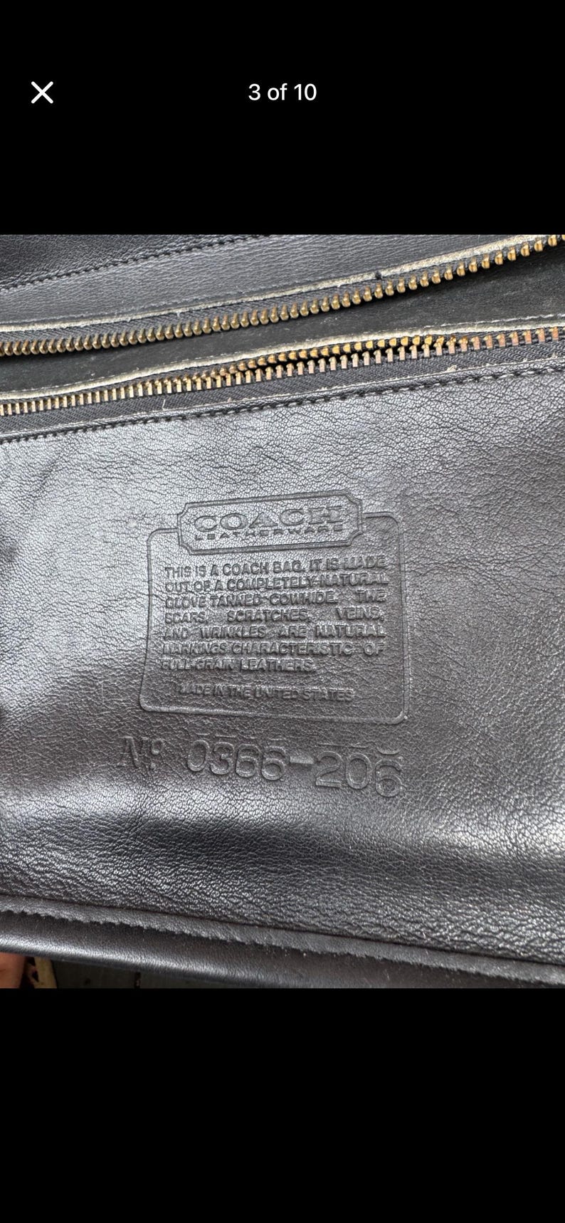 May include: Close-up of a black leather Coach bag. The bag has a textured surface with a gold zipper and a rectangular leather tag with text. The tag reads "Coach Leatherware" and includes text about the bag's materials and origin.