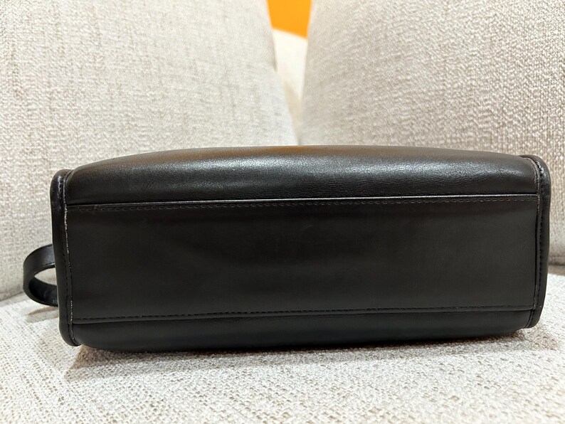 May include: A black leather handbag with a rectangular shape. The bag has a handle on one side and a smooth, shiny surface. The stitching is visible along the edges, and the bag appears to be well-constructed.