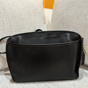 May include: A black leather crossbody bag with a rectangular shape. The bag has a front pocket and a long strap. The bag has a gold-colored zipper and a small leather tag attached to the side.