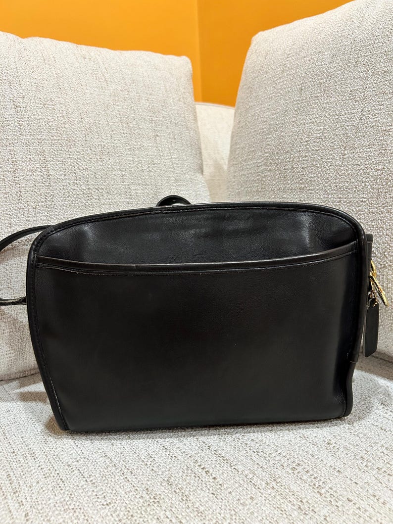 May include: A black leather crossbody bag with a rectangular shape. The bag has a front pocket and a long strap. The bag has a gold-colored zipper and a small leather tag attached to the side.