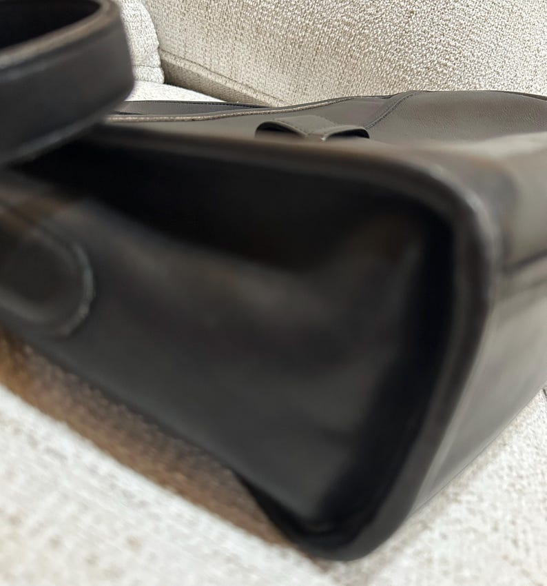 May include: Close-up of a black leather handbag with a shoulder strap. The bag has a rectangular shape with rounded corners and a subtle buckle detail on the top. The leather appears smooth and glossy.