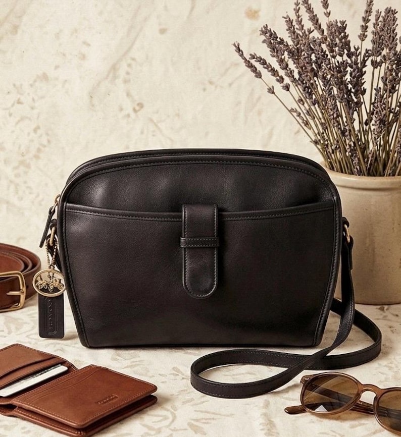 May include: A black leather crossbody bag with a flap closure and a long strap. The bag has a front pocket and gold-tone hardware. A brown leather wallet, belt, and sunglasses are also visible.