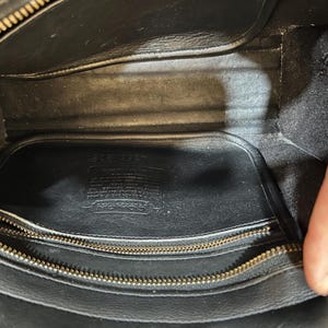 May include: Interior view of a black leather handbag. The bag features a gold zipper and a rectangular shape. The inside is lined with black fabric and includes a small pocket. The Coach logo is visible.