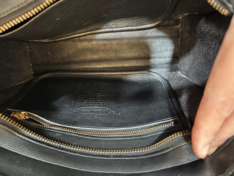 May include: Interior view of a black leather handbag. The bag features a gold zipper and a rectangular shape. The inside is lined with black fabric and includes a small pocket. The Coach logo is visible.