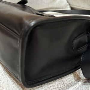 May include: A close-up of a black leather handbag. The bag has a rectangular shape with rounded corners and a shoulder strap. The leather appears smooth and glossy, with visible stitching along the edges.