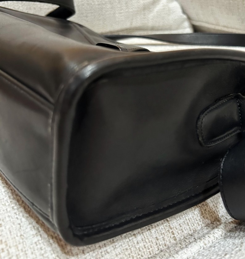 May include: A close-up of a black leather handbag. The bag has a rectangular shape with rounded corners and a shoulder strap. The leather appears smooth and glossy, with visible stitching along the edges.