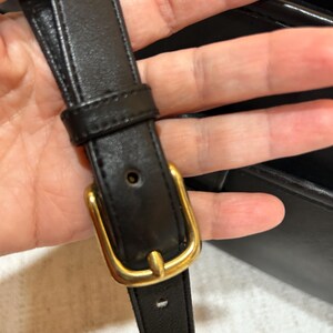 May include: Close-up of a black leather strap with a gold-colored buckle. The strap is held in place by a gold-colored buckle. The leather appears to be smooth and the buckle is rectangular.