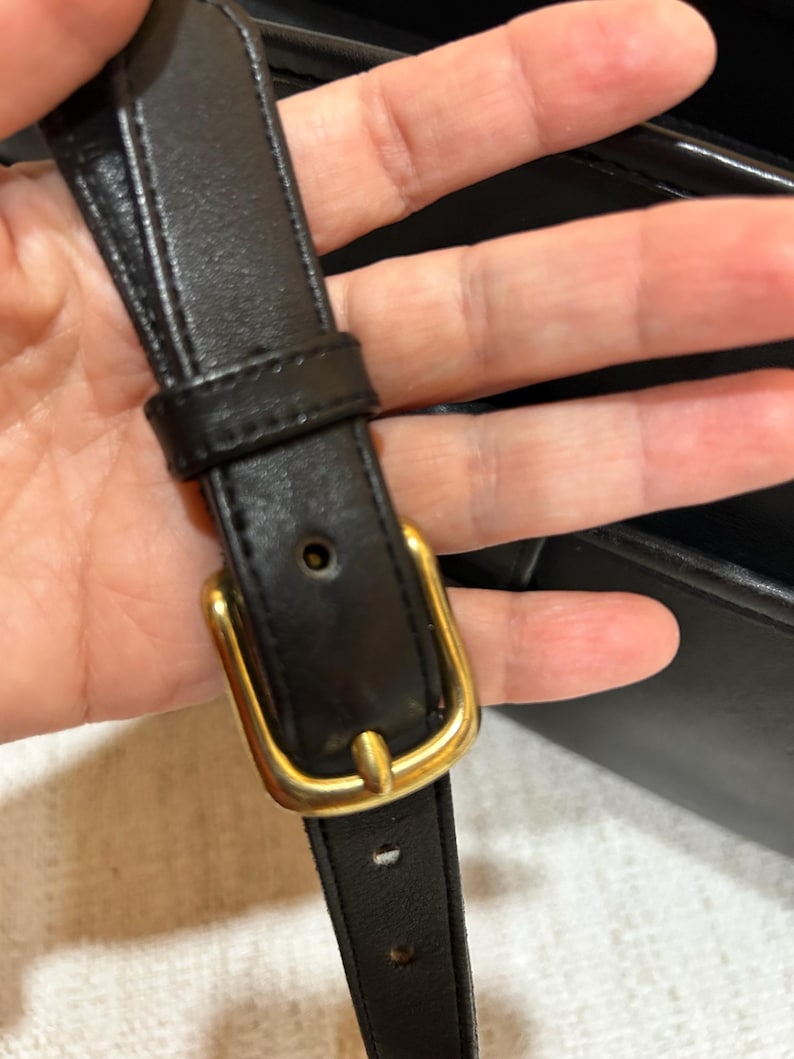 May include: Close-up of a black leather strap with a gold-colored buckle. The strap is held in place by a gold-colored buckle. The leather appears to be smooth and the buckle is rectangular.