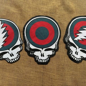 May include: Three skull-shaped patches with a white outline and black details. Each patch features a teal circle with a red bullseye. Two patches have a white lightning bolt, and one has a solid red circle.