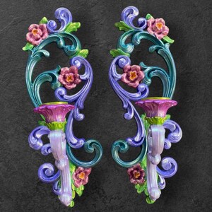 May include: A pair of ornate wall sconces with a floral design. The sconces are painted in shades of purple, teal, and green. Each sconce features a candle holder and a floral motif.
