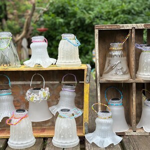 May include: A collection of twelve glass lamp shades repurposed as solar lanterns, each featuring a unique beaded wire handle. The shades vary in texture, including frosted, clear, and embossed patterns, and are displayed in two wooden crates.