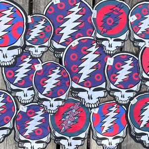 May include: A collection of circular fabric patches featuring a white skull graphic with a lightning bolt design. The background of each patch displays a pattern of red rings on a blue or purple field, with small white paper labels marked A through O.