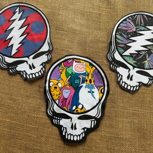 May include: Three skull-shaped patches with white teeth and black outlines. One patch features a red, blue, and white design, another has an Adventure Time cartoon scene, and the third displays a colorful cannabis leaf pattern.