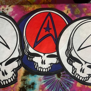 Op de afbeelding: Drie ronde patches met een schedelontwerp en een ster- en pijlsymbool. Eén patch heeft een rood midden met een blauw symbool, de andere hebben witte middens en zwarte symbolen. Op een kleurrijke tie-dye achtergrond.
