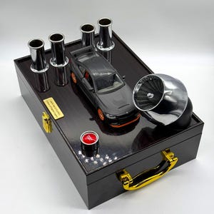 Op de afbeelding: Een zwarte houten kist met een donkerbruin interieur, met een grijze miniatuur sportwagen met oranje accenten, verchroomde uitlaatpijpen en een turbine. De kist heeft een gouden handvat en gouden accenten.