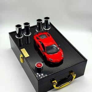 Op de afbeelding: Een zwarte houten kist met een glanzende afwerking, met een rode speelgoedauto en vier zilveren buizen. De kist heeft een gouden handvat en een kleine rode knop. Een gouden plaatje is aan de kist bevestigd.