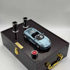 以下が含まれることがあります： 濃い木製の箱に、水色のミニカー、2つの黒い円筒、赤いボタンが置かれています。 箱には金色の金具と小さな金の銘板が付いています。 車はコンバーチブルです。