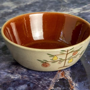 May include: A ceramic bowl with a cream-colored exterior and a glossy, reddish-brown interior. The bowl features a hand-painted design of a stylized tree with green leaves and yellow and red fruit. The bowl is perfect for serving food.