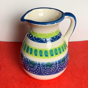 May include: A ceramic pitcher with a cream-colored base, adorned with blue, green, and white painted designs. The pitcher has a curved handle and a spout, with a blue rim. The decorative bands feature oval and dot patterns.