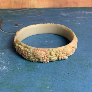 May include: Ivory-colored bangle bracelet with carved floral design. The bracelet features detailed relief carvings of flowers and leaves, with subtle pink accents. The bracelet is a vintage piece, likely made of resin or a similar material.