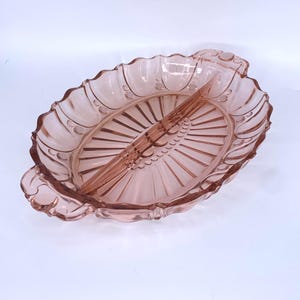 May include: A vintage pink glass divided serving dish. The oval-shaped dish features a scalloped edge and a central divider. The dish has a detailed design with a fan-like pattern in the center and handles on each end. A decorative piece for serving snacks.