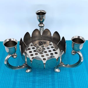 May include: Silver-toned metal centerpiece with a floral design. The piece features a central flower-shaped bowl with multiple holes, flanked by two candle holders on curved arms, and a single candle holder rising from the center.
