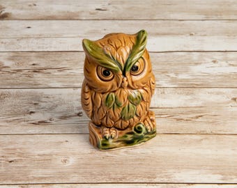May include: A ceramic owl figurine with brown and green details. The owl has large, round eyes and is perched on a green-painted log base. The figurine is approximately 6 inches tall.