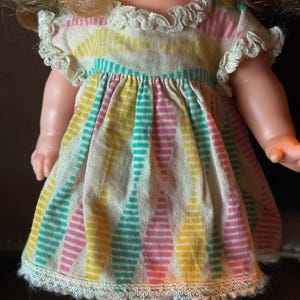 May include: A vintage doll wearing a colorful dress. The dress features vertical stripes in yellow, pink, and teal on a cream background. The dress has a ruffled collar and sleeves, and a lace trim at the hem. The doll has blonde hair and rosy cheeks.