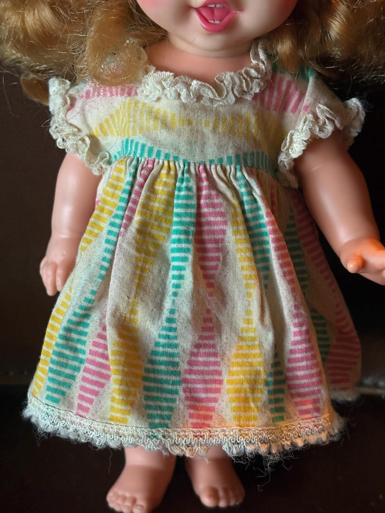 May include: A vintage doll wearing a colorful dress. The dress features vertical stripes in yellow, pink, and teal on a cream background. The dress has a ruffled collar and sleeves, and a lace trim at the hem. The doll has blonde hair and rosy cheeks.