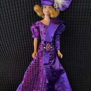 Puede incluir: Un vestido morado de lentejuelas y satén con un sombrero morado y una pluma. La muñeca tiene el pelo rubio y los ojos azules.