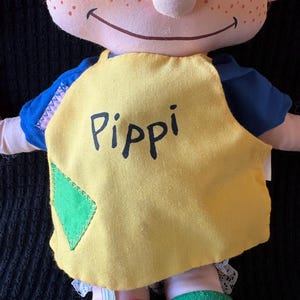 May include: A handmade doll with a smiling face, red hair, and freckles. The doll wears a yellow top with the name "Pippi" printed on it, blue sleeves, and mismatched red and green socks. A green diamond patch is sewn on the yellow top.