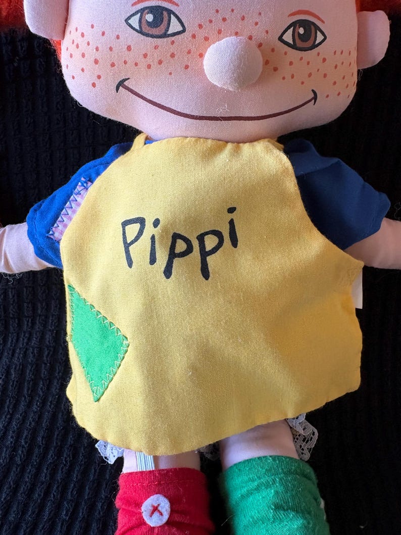 May include: A handmade doll with a smiling face, red hair, and freckles. The doll wears a yellow top with the name "Pippi" printed on it, blue sleeves, and mismatched red and green socks. A green diamond patch is sewn on the yellow top.