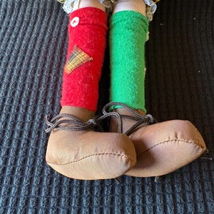May include: A doll's legs and feet are shown, with red and green socks and brown shoes. The red sock has a yellow and brown patch and a white button. The shoes have brown laces.