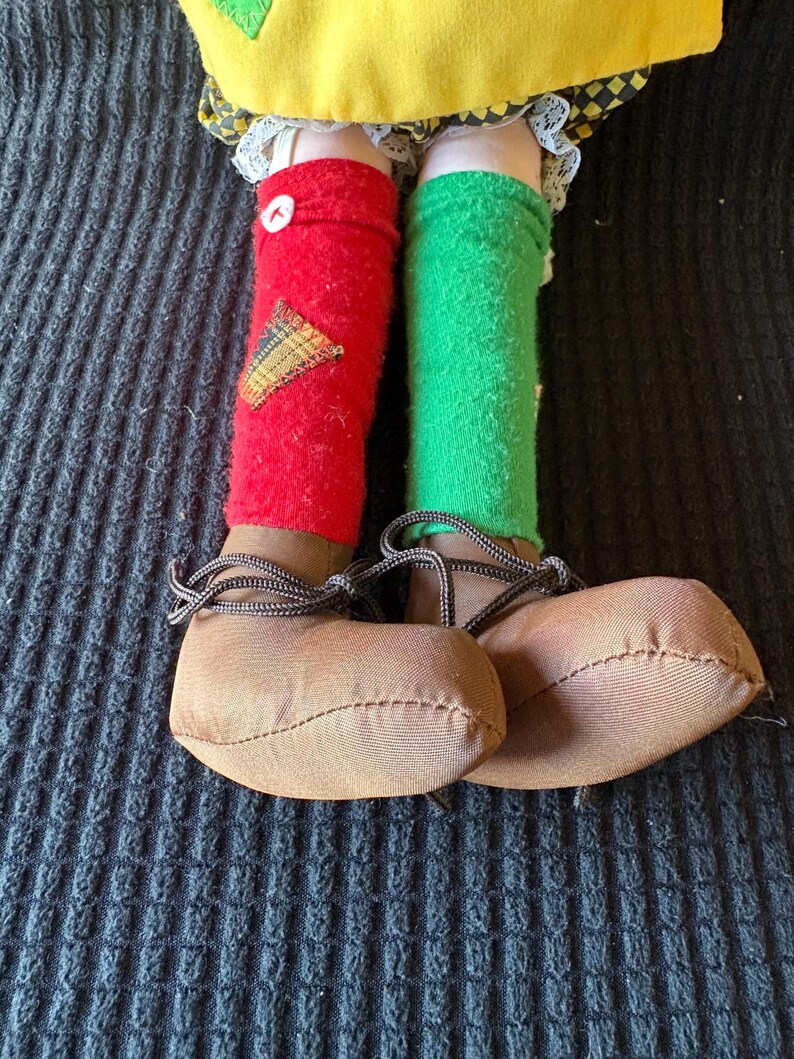 May include: A doll's legs and feet are shown, with red and green socks and brown shoes. The red sock has a yellow and brown patch and a white button. The shoes have brown laces.