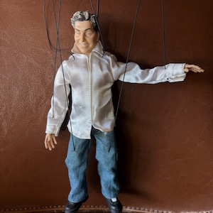 Może przedstawiać: Marionetka ubrana w białą satynową koszulę, niebieskie dżinsy i czarne buty. Lalka ma uśmiechniętą twarz i szare włosy. Czarne sznurki są przymocowane do manipulacji.