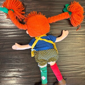 May include: A handmade doll with bright orange yarn pigtails tied with green ribbons. The doll wears a blue top, yellow braces, checkered shorts, mismatched socks, and brown shoes. The doll is lying down.