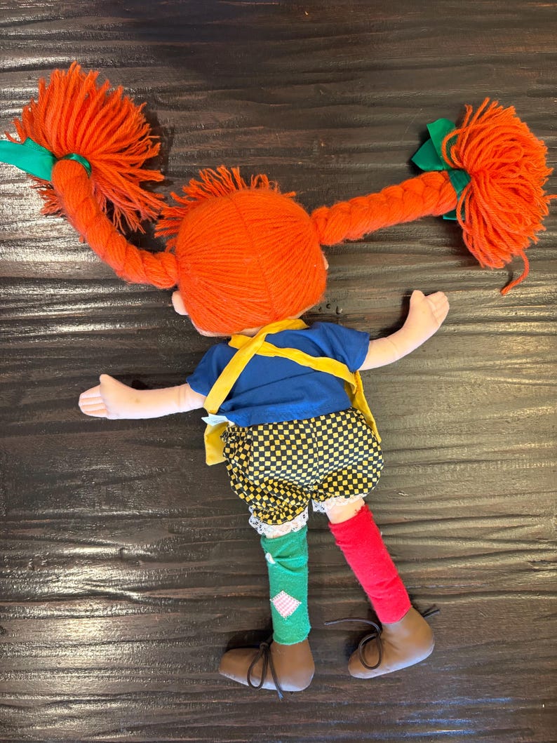 May include: A handmade doll with bright orange yarn pigtails tied with green ribbons. The doll wears a blue top, yellow braces, checkered shorts, mismatched socks, and brown shoes. The doll is lying down.
