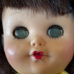 May include: Close-up of a vintage doll's face. The doll has dark brown hair, green eyes, and red lips. The doll's cheeks are blushed, and it is wearing a yellow top. The doll's face is made of a smooth material.