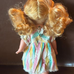 May include: A vintage doll with golden blonde hair styled in pigtails. The doll wears a colorful striped dress with pink, yellow, and teal vertical stripes. The dress has a white lace trim around the sleeves and hem.