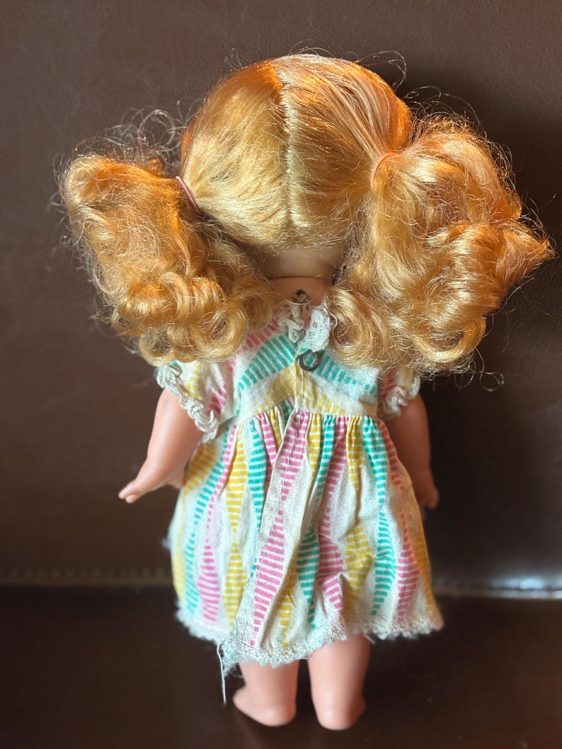 May include: A vintage doll with golden blonde hair styled in pigtails. The doll wears a colorful striped dress with pink, yellow, and teal vertical stripes. The dress has a white lace trim around the sleeves and hem.