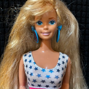 May include: A blonde doll with blue eyes and a white and blue star patterned dress. The doll is wearing blue hoop earrings.