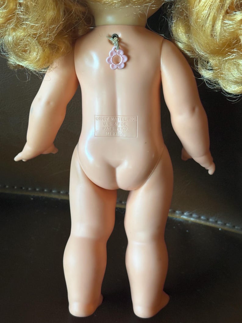 May include: Back view of a vintage doll with blonde hair. The doll has a light skin tone and is wearing a pink flower charm. The text on the doll reads "&copy;1967 Mattel Inc. U.S.A. For Pat. Pend. Mexico."