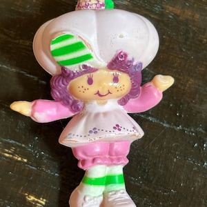 May include: A vintage plastic doll with purple hair, a pink dress, and a white hat with green stripes. The doll is wearing pink shoes with green stripes and has a smiling face.