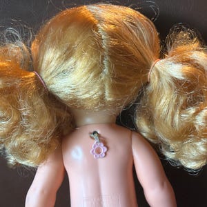 May include: A vintage doll with blonde hair styled in pigtails, secured with pink rubber bands. The doll's back is visible, featuring a pink flower-shaped charm. The doll's skin is a light peach color.