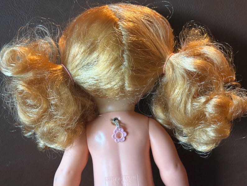 May include: A vintage doll with blonde hair styled in pigtails, secured with pink rubber bands. The doll's back is visible, featuring a pink flower-shaped charm. The doll's skin is a light peach color.