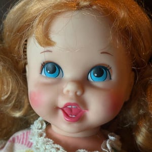 May include: Close-up of a vintage doll with blonde hair and blue eyes. The doll has rosy cheeks and a pink mouth. It is wearing a dress with a white lace collar and a striped pattern in pastel colors.