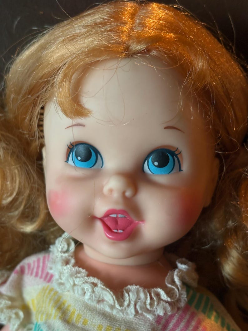 May include: Close-up of a vintage doll with blonde hair and blue eyes. The doll has rosy cheeks and a pink mouth. It is wearing a dress with a white lace collar and a striped pattern in pastel colors.