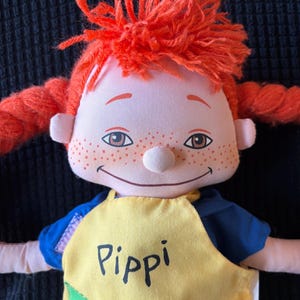 May include: A soft doll with bright red yarn hair in braids and a top knot. The doll has a smiling face, freckles, and is wearing a yellow top with the name "Pippi" printed on it. The doll's shirt has blue sleeves.